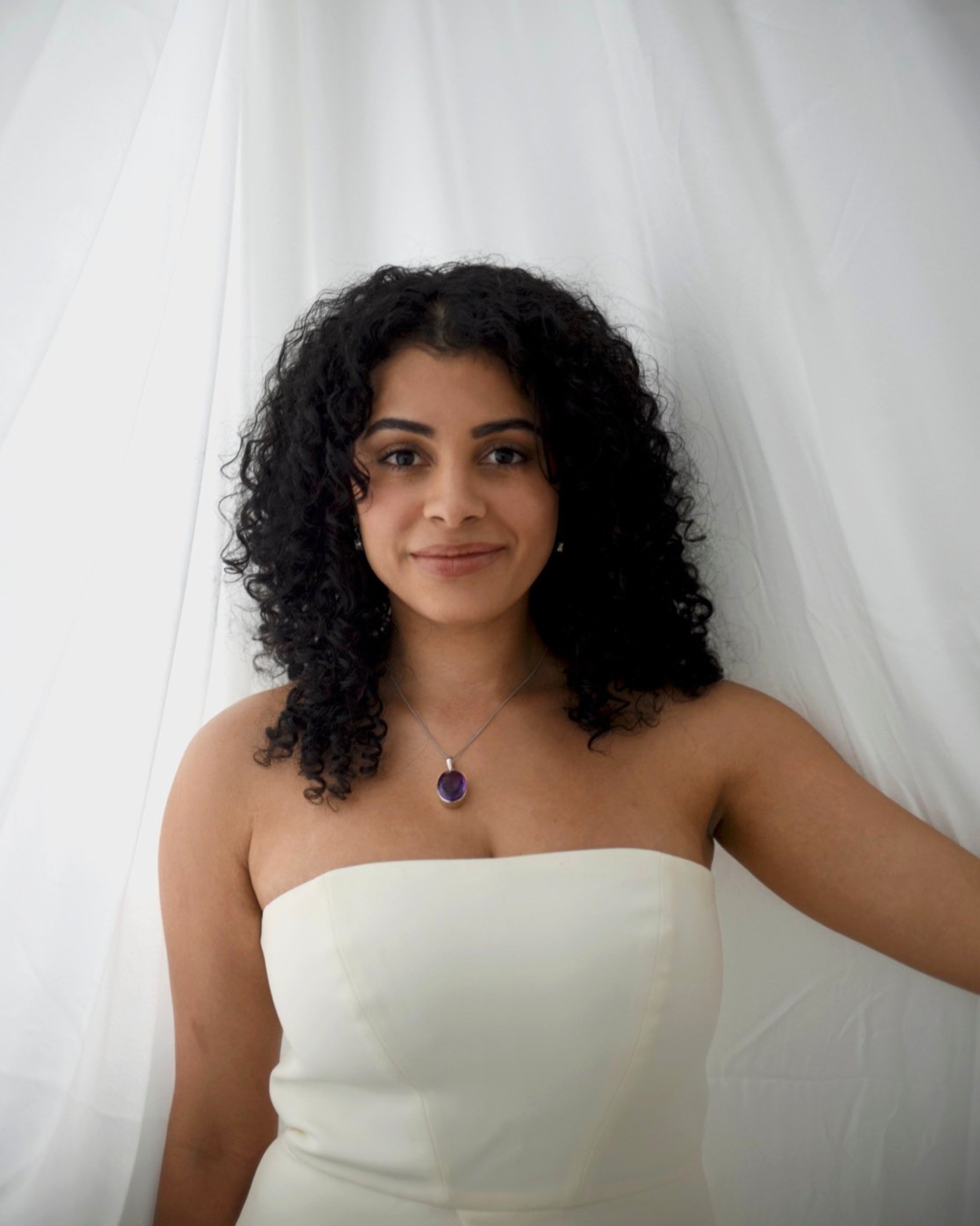 A woman with curly black hair wearing a strapless white dress and a purple pendant necklace, posing in front of a white draped fabric backdrop.