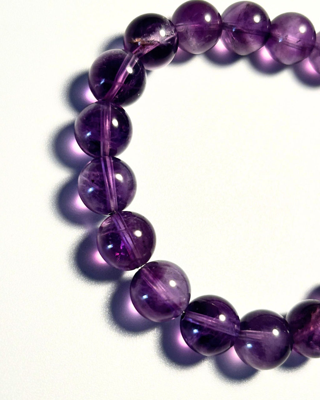 A close-up of a beaded bracelet made of polished purple translucent gemstone beads, arranged in a curved circle on a white background with soft shadows.