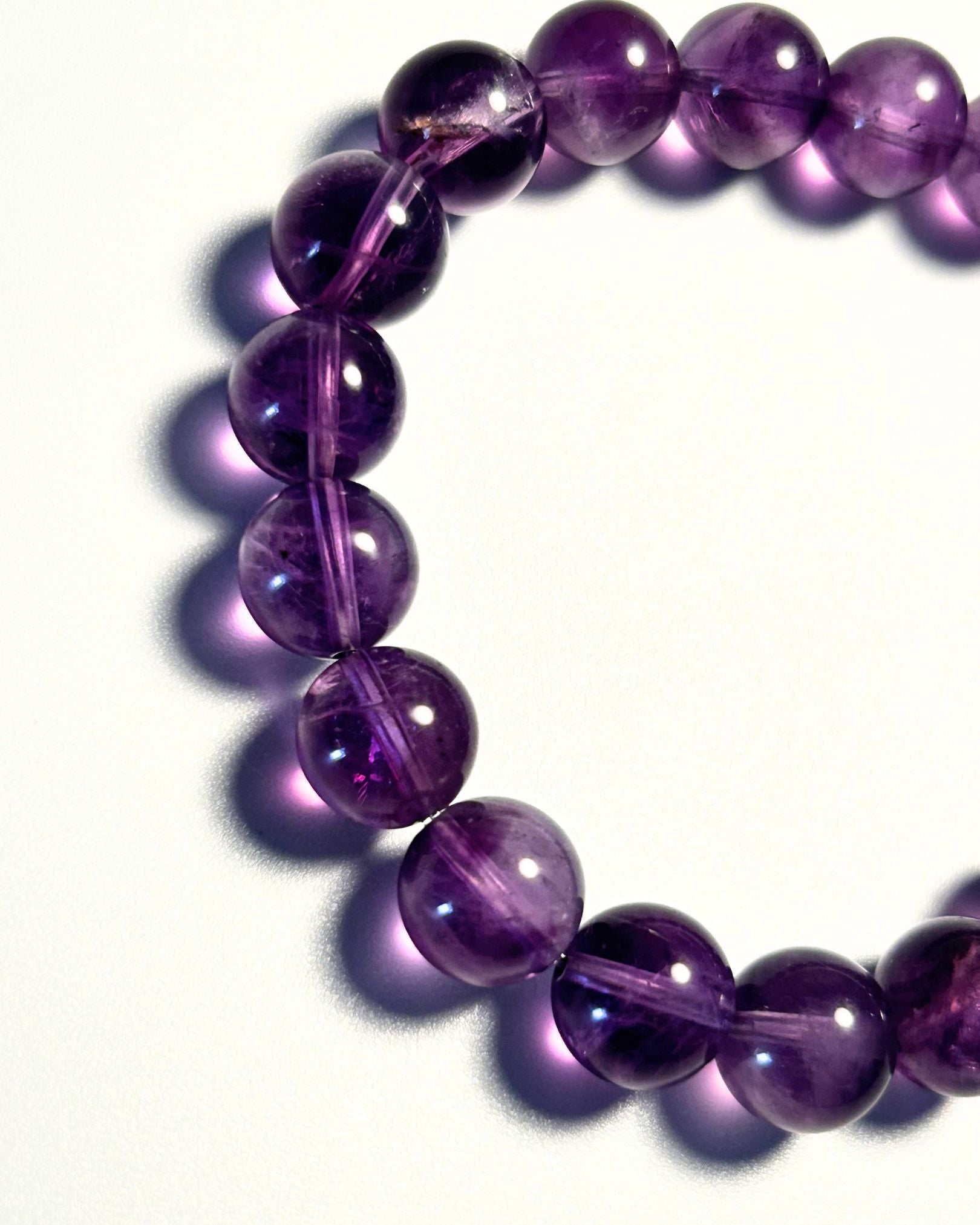 A close-up of a beaded bracelet made of polished purple translucent gemstone beads, arranged in a curved circle on a white background with soft shadows.