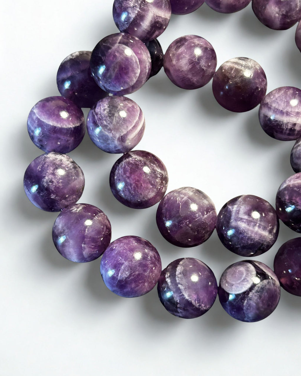 A close-up of a beaded bracelet made of polished round purple gemstone beads with natural banding and iridescent highlights, arranged on a plain light background.
