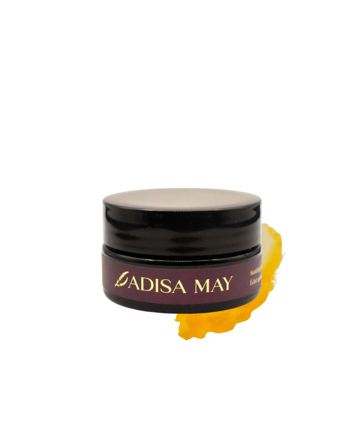 A small black cosmetic jar with a dark mauve label reading 'ADISA MAY' in gold lettering, shown on a white background with a yellow cream smear behind it.