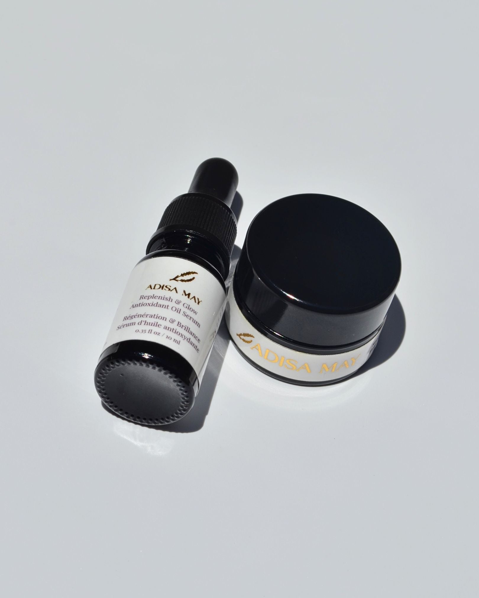 A small black dropper bottle and a matching black jar with white labels and gold lettering, photographed on a clean light gray background.
