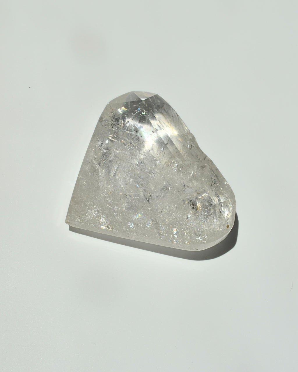 A raw, faceted clear quartz crystal stone with visible internal inclusions and a triangular shape, resting on a plain light gray surface with a soft shadow.
