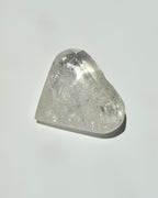 A raw, faceted clear quartz crystal stone with visible internal inclusions and a triangular shape, resting on a plain light gray surface with a soft shadow.