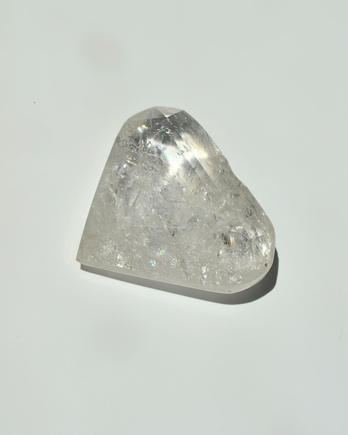 A raw, faceted clear quartz crystal stone with visible internal inclusions and a triangular shape, resting on a plain light gray surface with a soft shadow.