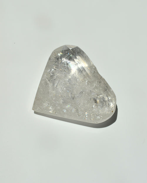 A raw, faceted clear quartz crystal stone with visible internal inclusions and a triangular shape, resting on a plain light gray surface with a soft shadow.
