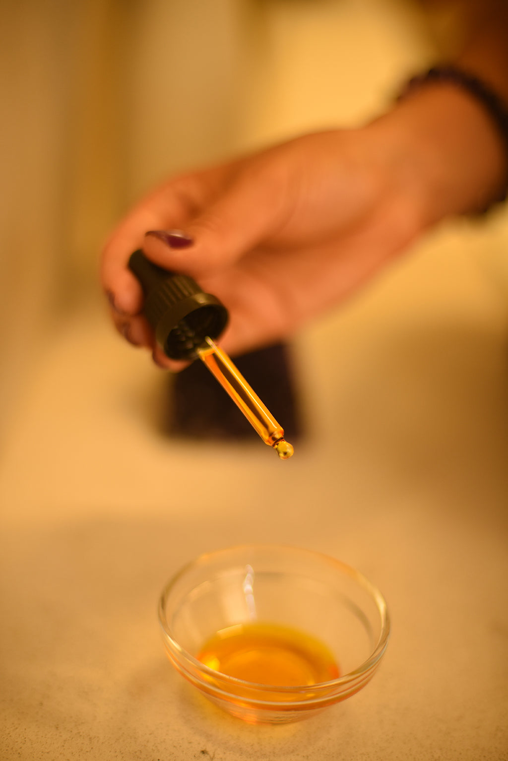 Hand holding a dropper over a small glass bowl with a yellow liquid on a blurred background
