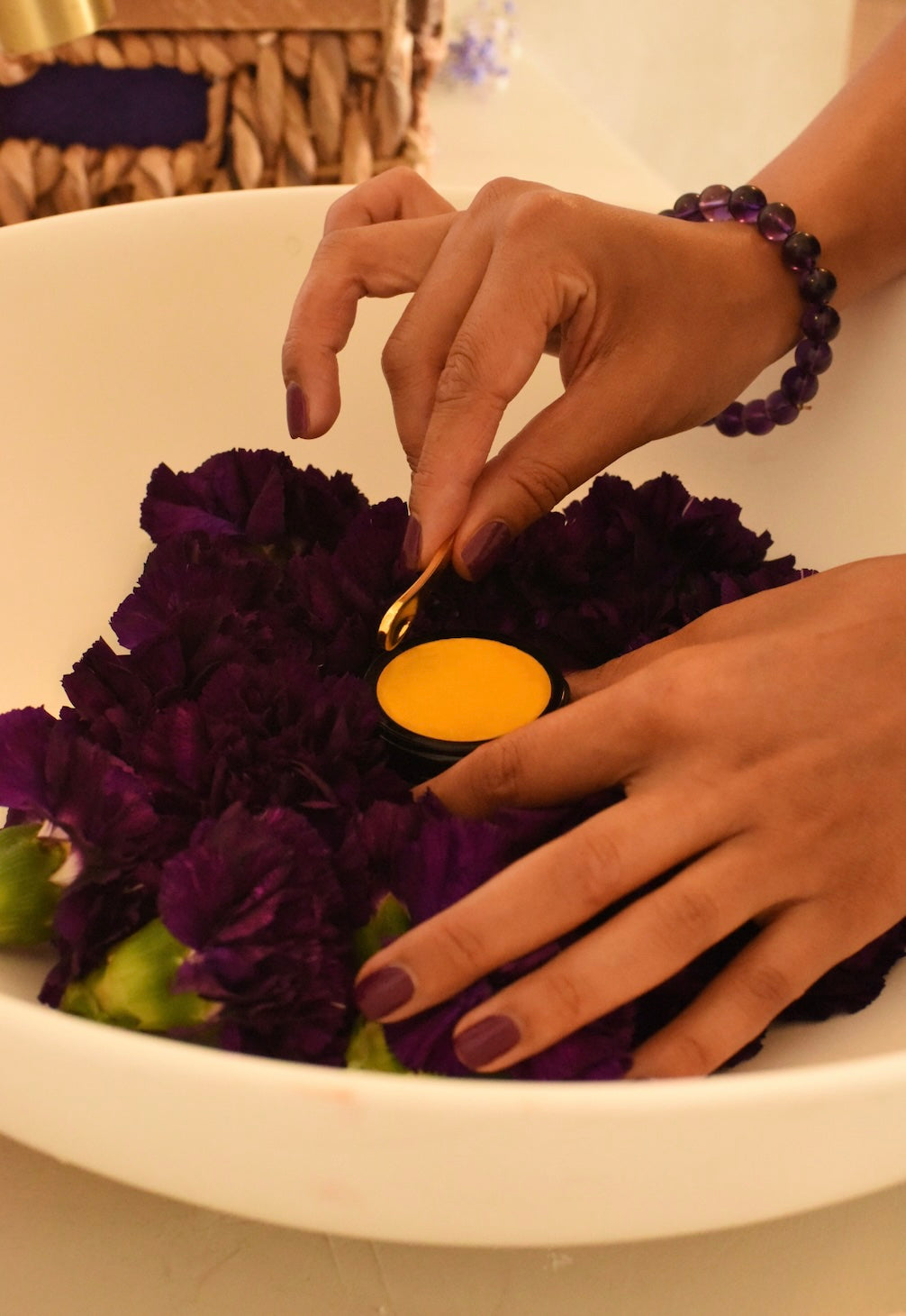 Person placing a golden coloured cosmetic balm for dry skin among purple flowers in a white bowl.